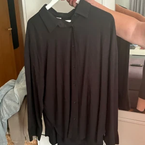 Svart långärmad skjorta med knappar - Stilren svart skjorta med klassisk krage och knäppning framtill. Skjortan har lång ärm och en loose passform som ger en chill vibe. Perfekt att styla med jeans eller över en t-shirt för en avslappnad look.