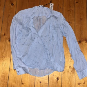 Ljusblå omlottblus från Angelology - Säljer en ljusblå, transparent omlottblus från Angelology i storlek S. Blusen har lång ärm, v-ringad hals och är gjord i ett tunt, flowy material som ger en chill vibe. Perfekt för dig som gillar stilrena och luftiga toppar med lite extra edge.