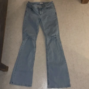 Bootcut ljusblå jeans med slitningar - Ljusblå bootcut jeans med slitningar över knäna och snygga detaljer på bakfickorna i form av röda och vita sömmar. Klassisk femficksmodell i jeansmaterial, perfekt för dig som gillar en retroinspirerad look med lite extra edge.