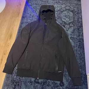 Bluewear svart jacka, storlek xs - Svart vindjacka fr med huva och snörning. Jackan har flera dragkedjeförsedda fickor fram och på ärmen, samt ribbade muddar vid ärmslut och nederkant. Perfekt för dig som gillar stilren och funktionell design.