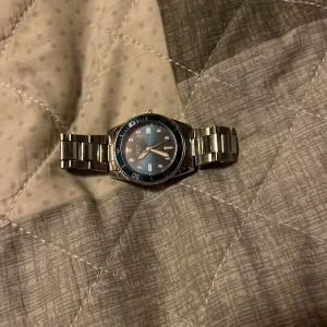 Blå och silverfärgad klocka från Regal - Säljer en snygg klocka från Regal med blå urtavla och silverfärgad boett och länkarmband i metall. Klockan har tydliga markeringar och en vridbar ring runt urtavlan. Extra länkar till armbandet medföljer så du kan justera storleken enkelt.