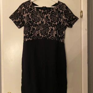 Svart spetsklänning med mönster - Snygg svart kortklänning med överdel i spets och coolt virvlande mönster. Klänningen har rund hals, korta ärmar och en slät svart nederdel. Spetsen är svart med ljus bakgrund som ger en kontrast. Perfekt för dig som gillar detaljer och stilren look.