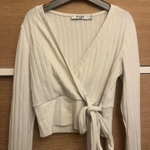 Vit omlottopp med knyt från NA-KD - Säljer en vit omlottopp från NA-KD i ribbad bomull. Toppen har långa ärmar, v-ringning och snygg knytning i midjan. Perfekt för dig som gillar stilrena och trendiga plagg med lite extra detalj. Passar dig som vill ha en croppad look.