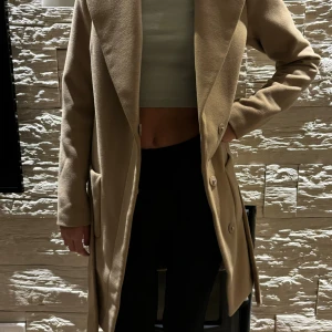 Beige kappa från Vila med bälte XS - Snygg beige kappa från Vila i storlek XS. Jackan har klassisk krage, knappar framtill, fickor på sidorna och ett bälte i midjan för en clean och stilren look. Perfekt för dig som gillar minimalistisk stil och vill ha en jacka som funkar till allt.