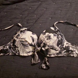En svart/vit blommig bikini! - Superfin svartvit bikinitopp med blommigt mönster. Har fasta axelband och en liten rosett framtill som gör den extra söt. Lätt vadderad och sitter bekvämt utan att glida. Perfekt till sommaren! I väldigt fint skick.                            Skriv vid intresse!🤍🖤🤍🖤                                          Jag vet tyvärr inte märket eller varifrån den är!