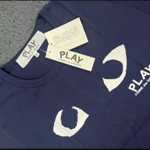 Mörkblå Comme des Garçons PLAY t-shirt - Säljer en mörkblå t-shirt från Comme des Garçons PLAY i storlek S. T-shirten har korta ärmar och är gjord i mjuk bomull. På framsidan finns ett vitt tryck med ögon och PLAY-loggan. Perfekt för dig som gillar streetwear och unik design.