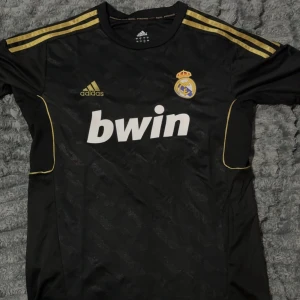 Real Madrid Kaka svart matchtröja - Säljer en svart Real Madrid fotbollströja från Adidas med guldiga detaljer och Kaka nummer 8 på ryggen. Tröjan har korta ärmar, klubbmärke på bröstet och Bwin-tryck framtill. Materialet är lätt och andas, perfekt för fotboll eller samling.