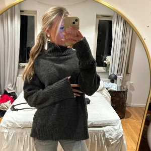 Mörkgrå stickad polotröja - Mysig mörkgrå stickad polotröja med oversized passform och långa ärmar. Tröjan har bred ribbstickad mudd vid ärmslut, nederkant och hals. Perfekt för kalla dagar och enkel att styla med jeans eller kjol.