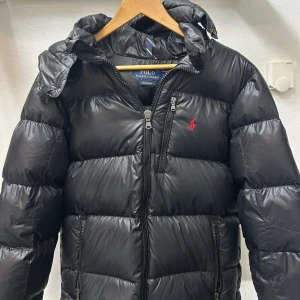 Svart pufferjacka Polo Ralph Lauren S - Svart pufferjacka från Polo Ralph Lauren med röd broderad logga på bröstet. Jackan har huva, flera dragkedjeförsedda fickor och RL-detaljer på dragkedjorna. Tillverkad i glansig polyester och perfekt för kalla dagar.