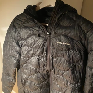 Svart dunjacka Peak Performance - Svart dunjacka från Peak Performance med vågformat quiltat mönster. Jackan har huva, hel dragkedja framtill och diskret logga på bröstet. Perfekt för kyliga dagar och riktigt skön passform.