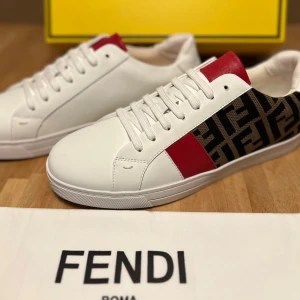 Fendi vita sneakers med röd detalj - Snygga vita sneakers från Fendi med röd läderdetalj på plösen och ikoniskt Fendi-mönster i svart och brunt på sidan. Skorna har rund tå, platt sula och klassisk snörning. Tillverkade i skinn för en lyxig känsla och stilren look.