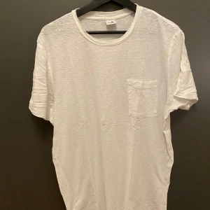 Vit t-shirt med ficka från NN07 - Vit t-shirt från NN07 i storlek M, tillverkad i mjuk bomull. Klassisk rund hals och en enkel bröstficka på vänster sida. T-shirten har korta ärmar och en clean, stilren look som funkar till allt. Perfekt för dig som gillar minimalistisk stil.