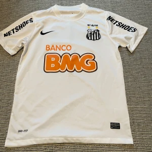 Vit Santos FC Nike fotbollströja - Santos FC matchtröja i vitt från Nike med klubbmärke, sponsorer som BMG och Netshoes samt Dri-FIT-material. Klassisk passform med rund hals och korta ärmar. Perfekt för dig som gillar brasiliansk fotboll och snygg sportstil.