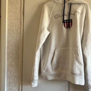 Vit hoodie från Gant med broderad logo - Vit hoodie från Gant med broderad logo och sköld på bröstet. Tröjan har huva med svarta snören, känguruficka och långa ärmar. Perfekt för en chill och sportig look. Använd 1-3 gånger max