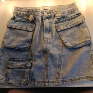 Jeanskjol med stora fickor från Yoyo S. - Snygg ljusblå jeanskjol från Yoyo S. med flera stora fickor framtill och baktill. Kjolen har en klassisk jeanslook med synliga sömmar, bälteshällor och knappstängning. Perfekt för dig som gillar praktiska detaljer och streetstil. STORLEK 10 ÅRINGAR