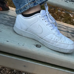 Vita Nike sneakers i skinn - Säljer ett par klassiska vita Nike sneakers i skinn med rund tå och snörning. Skorna har perforerade detaljer framtill för extra ventilation och en platt sula. Perfekta för dig som gillar en clean och tidlös look.