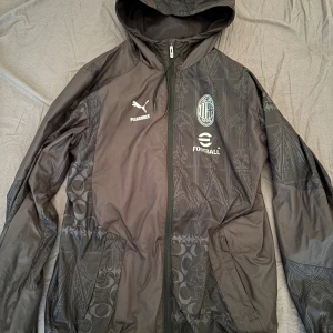 Svart AC Milan vindjacka Puma - Svart vindjacka från Puma x Pleasures med AC Milan-logga och eFootball-tryck. Jackan har huva, hel dragkedja och diskret mönster i tyget. Perfekt för fotbollsfans som vill ha en snygg och sportig look.