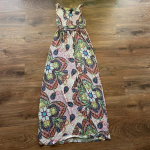 Färgglad långklänning  - Säljer en maxiklänning från MNG Casual Sportswear i storlek M. Klänningen har ett färgstarkt blommigt och paisley-inspirerat mönster i blått, rött, grönt och gult. Ärmlös modell med rund hals och markerad midja. Perfekt för dig som gillar bohemisk stil och vill sticka ut.