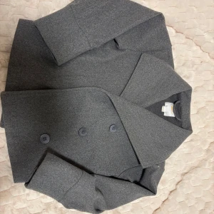 Grå rock från Gina Tricot - Snygg grå rock från Gina Tricot med bred krage och dubbelknäppning framtill. Jackan har långa ärmar och en stilren, rak passform. Perfekt för dig som gillar klassisk och enkel design med en modern touch.