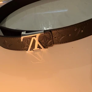 Bälte  - Snyggt svart bälte  skinn med klassiskt monogram präglat längs hela remmen. Bältet har en stilren, silverfärgad  som ger en lyxig känsla. Perfekt accessoar för att lyfta din outfit med en exklusiv touch.