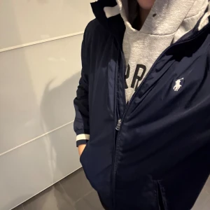 Marinblå vindjacka från Polo Ralph Lauren - Snygg marinblå vindjacka från Polo Ralph Lauren med vit broderad logga på bröstet. Jackan har dragkedja, två fickor med dragkedja och ribbade muddar med vit detalj. Perfekt för dig som vill ha en stilren och sportig look.