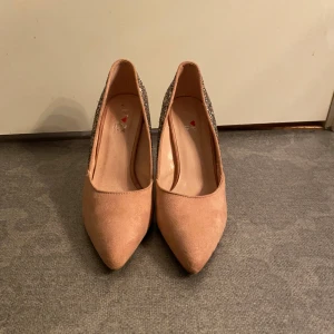 Glittriga pumps med spetsig tå - Snygga pumps i beige mockaliknande material framtill och med glittrig baksida och klack. Skorna har spetsig tå och hög klack, perfekta för att sticka ut. Glittret på baksidan ger en extra festlig touch.Passar perfekt för en ute kväll med kompisarna 🥳🎉🪩