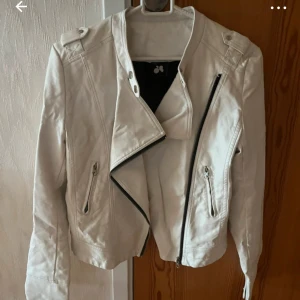 Vit skinnjacka med dragkedja - Snygg vit skinnjacka med asymmetrisk dragkedja och coola detaljer på axlarna. Jackan har två fickor med dragkedja och en klassisk bikerstil.😊