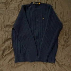 Mörkblå stickad tröja Polo Ralph Lauren - Säljer en mörkblå stickad tröja från Polo Ralph Lauren med klassiskt broderat logotyp på bröstet. Tröjan har rund halsringning, ribbade muddar och ett stilrent, ribbat mönster. Perfekt för dig som gillar en clean och tidlös look. Den är L men sitter som S!
