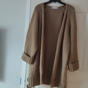 Beige stickad kofta från Zara - Mysig beige stickad kofta från Zara med lång ärm och uppvikta muddar. Koftan är öppen framtill och har en chunky stickad struktur som ger en avslappnad och trendig look. Perfekt att slänga på sig när det är kyligt ute.