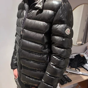Svart Moncler dunjacka herr - Säljer en svart, glansig dunjacka från Moncler med klassisk logga på ärmen. Jackan har quiltad design, dragkedja framtill och hög krage med knappdetalj. Perfekt för kalla dagar och riktigt snygg till streetwear-stil.