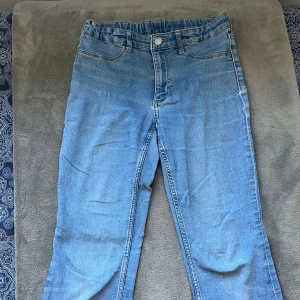 Ljusblå jeans från H&M, flat leg - Säljer ett par ljusblå jeans från H&M med flat leg och justerbar midja. Klassisk femficksmodell med låg midja och knappstängning. Perfekta för dig som gillar en avslappnad och enkel stil.