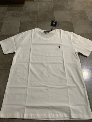 Vit t-shirt från Polo Ralph Lauren XL - Vit t-shirt från Polo Ralph Lauren i storlek XL. Klassisk design med rund hals och korta ärmar. T-shirten är gjord i mjuk bomull och har den ikoniska broderade Polo-loggan på bröstet. Perfekt för en clean och stilren look.