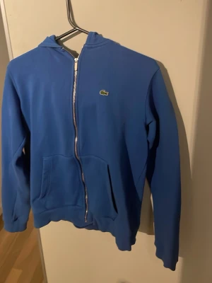 Blå Lacoste hoodie - En stilren blå Lacoste zip hoodie i mjuk bomull med den ikoniska krokodilen på bröstet. Perfekt balans mellan sportig elegans och vardagskomfort, ett självklart val för en snygg avslappnad look👀 