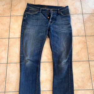 Nudie jeans  - Snygga blå jeans från Nudie Jeans med klassisk femficksdesign och orangea sömmar. Jeansen har raka ben och normal passform, med tydliga slitningar för en cool look. Materialet är robust denim i bomull och de har knappgylf framtill. Litet hål mellan fick sömmen men inget man märker och väldigt enkelt att laga!  PRIS KAN DISKUTERAS 