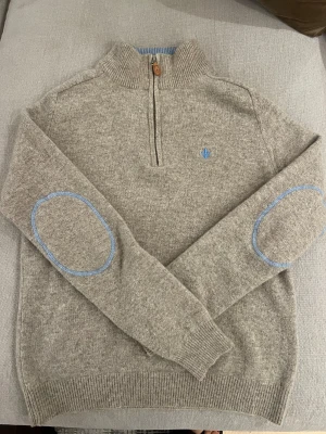 Grå stickad half zip-tröja Morris - Snygg grå stickad tröja från Morris med half zip och ribbad krage i ljusblått. Broderad blå logga på bröstet och matchande blå detaljer på armbågarna. Perfekt för dig som gillar klassisk stil med en twist. Tröjan är liten i storlek.