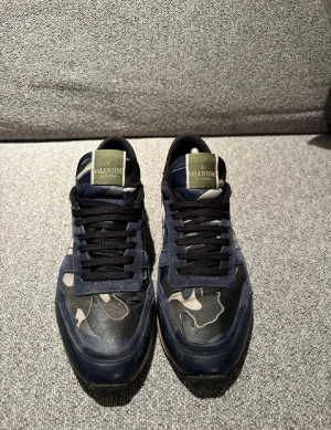 Valentino Garavani Rockrunner sneakers - Snygga Valentino Garavani Rockrunner sneakers i mörkblått och grått med camouflage-mönster. Skorna har svarta snören, rund tå och detaljer i mocka och skinn. Klassisk logga på plösen och en liten svart sula. Perfekta för dig som vill sticka ut med stil.