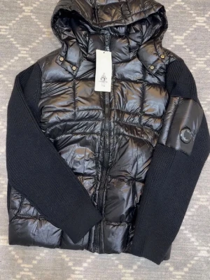 Svart dunjacka från C.P. Company - Svart dunjacka från C.P. Company med quiltad front och huva, stickade ärmar och coolt linsmärke på vänster arm. Jackan har dragkedja och ribbade muddar, perfekt för dig som gillar streetwear med edge. Insidan är fodrad och har märkets patchar.