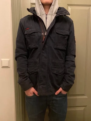 Svart fieldjacka från Superdry - En riktigt snygg svart fieldacka från Superdry med fyra stora fickor framtill, hög krage och coola detaljer som patch på ärmen och röda sömmar. Jackan har dragkedja och knappar framtill. Vid funderingar, fråga på! 