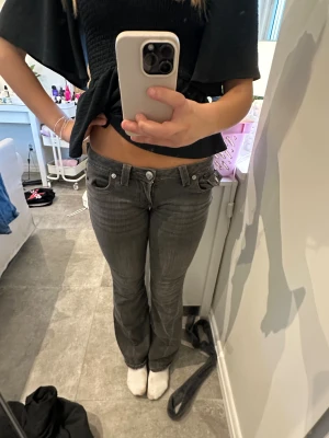 Gråa lågmidjade bootcut jeans - Säljer ett par gråa true religion lågmidjade jeans med bootcut passform. Jeansen har snygga detaljer som stora bakfickor med lock och silverfärgade knappar. Perfekta för dig som gillar en avslappnad men trendig look.