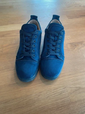 Loubutin skor - Snygga marinblå sneakers från Christian Louboutin i mjuk mocka med klassisk snörning och rund tå. Skorna har en röd sula med Louboutin-logga och broderad text på hälen. Insidan är beige och sulan är platt. Perfekt för dig som gillar exklusiva detaljer.