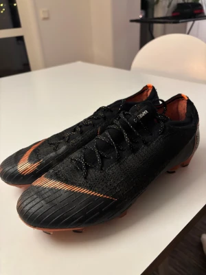 Nike Mercurial Vapor 13 fotbollsskor - Säljer ett par svarta limiterade Nike Mercurial Vapor 13 fotbollsskor med orange detaljer och logga. Skorna har snörning, platt sula med dobbar och är tillverkade i syntetmaterial med stickad ovandel för extra komfort och passform. Perfekta för dig som vill ha grym kontroll och speed på planen.