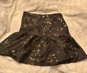 Svart och guld kjol Holly & Whyte - Supersnygg kort kjol från Holly & Whyte by Lindex i svart tyg med glittriga guldiga blommönster. Kjolen har en lätt utställd form och dragkedja i sidan. Perfekt för dig som vill sticka ut med metallic-detaljer och cool struktur. Perfekt skick 10/10. Passar på julfest, nyårsfest och till vardags beroende på vad man har till.