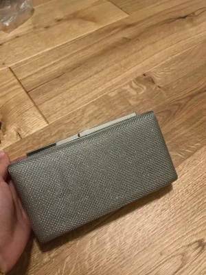 Silverfärgad kuvertväska med metallram - Stilren kuvertväska i silver med metallram upptill. Väskan har en rektangulär form och ett diskret glittrigt yttre som ger en lyxig känsla. Perfekt att bära i handen när du vill addera lite extra bling till din outfit.