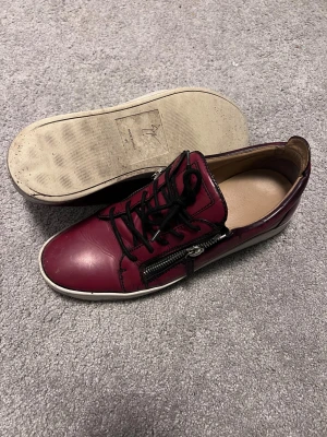 Vinröda sneakers från Giuseppe Zanotti - Snygga vinröda sneakers från Giuseppe Zanotti med svart snörning och dragkedja på sidan. Skorna har en rund tå och vit platt sula. Tillverkade i skinn med coola detaljer som ger en lyxig vibe. Perfekta för dig som gillar statement-skor.