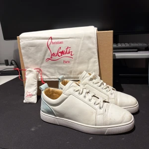 Christian Louboutin vita sneakers - Säljer ett par vita sneakers från Christian Louboutin i skinn med ikonisk röd sula och ljusblå detalj på hälen. Skorna har rund tå, snörning och diskret logga bak. Perfekta för dig som vill ha en clean men lyxig look med exklusiv känsla.