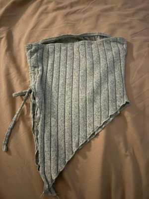 Grå ribbad tubtopp med snörning - Säljer en grå tubtopp med ribbad struktur och snörning på sidan. Toppen har en asymmetrisk, spetsig nederkant som ger en cool och unik look. Materialet är mjukt och stretchigt, perfekt för att styla med jeans eller mjukisbyxor. Sitter tight och är axelbandslös.
