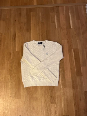 Vit kabelstickad tröja från Polo Ralph Lauren - Snygg vit kabelstickad tröja från Polo Ralph Lauren med klassisk marinblå broderad logga på bröstet. Tröjan har rund hals, långärmad passform och är tillverkad i mjuk bomull. Perfekt för dig som gillar stilrena och tidlösa plagg. Pris kan diskuteras.