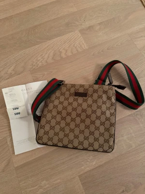 Gucci axelväska med GG-mönster - Snygg axelväska från Gucci i beige och brunt med klassiskt GG-mönster. Väskan har en bred axelrem i grönt och rött, samt dragkedja upptill. Materialet är canvas med detaljer i skinn. Perfekt storlek för det viktigaste och har en innerficka med dragkedja.