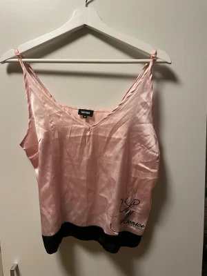 Rosa satinlinne från Bubbleroom - Snygg ljusrosa satinlinne  från Bubbleroom med svarta detaljer längst ner och broderad text 'Monroe' i svart. Tunn, glansig och mjuk i satinliknande material, med smala axelband och v-ringad hals. 