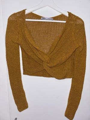 Senapsgul stickad omlott-tröja Zara - Säljer en senapsgul stickad tröja från Zara med omlott-detalj framtill och djup v-ringning. Tröjan är croppad och har långa ärmar, perfekt för att ge din outfit en cool touch. Materialet är grovstickat och mjukt.